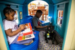 Two children read in a cozy blue nook; a black dog sits at their feet, listening attentively.