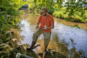 A photo of Andrew Shaugnassy standing on a riverbank. He holds a plastic sample bottle. 